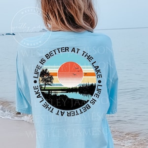 May include: A light blue t-shirt with a circular graphic design featuring a sunset over a lake with trees and the text "Life is better at the lake".
