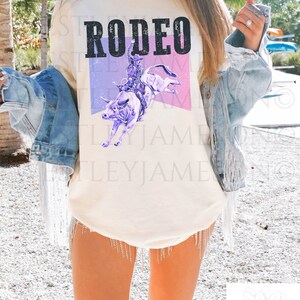 Puede incluir: Una camiseta blanca con la palabra "RODEO" impresa en letras negras. Un gr&aacute;fico morado y rosa de un vaquero montando un toro est&aacute; impreso debajo de la palabra. La camiseta la lleva puesta una persona. El texto "IVORY" est&aacute; impreso en la esquina inferior izquierda de la imagen. El texto "WESTLEY JAMESON" est&aacute; impreso en la esquina inferior derecha de la imagen.