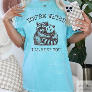 K&ouml;nnte beinhalten: Ein hellblaues T-Shirt mit einem Aufdruck eines Waschb&auml;ren und eines Opossums, die sich umarmen. Der Text "You're Weird I'll Keep You" ist auf dem Shirt gedruckt.