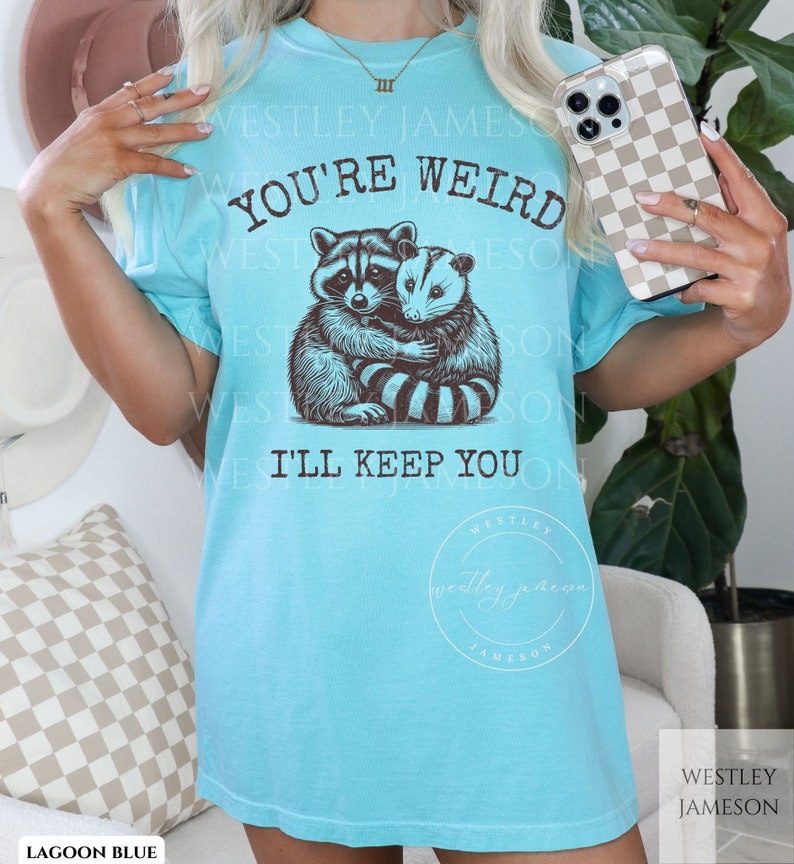 K&ouml;nnte beinhalten: Ein hellblaues T-Shirt mit einem Aufdruck eines Waschb&auml;ren und eines Opossums, die sich umarmen. Der Text "You're Weird I'll Keep You" ist auf dem Shirt gedruckt.