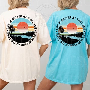 May include: Two t-shirts with a graphic design on the back. The design is a circle with a sunset, trees, and a lake. The text around the circle says "Life is better at the lake". The t-shirt on the left is ivory and the t-shirt on the right is light blue.