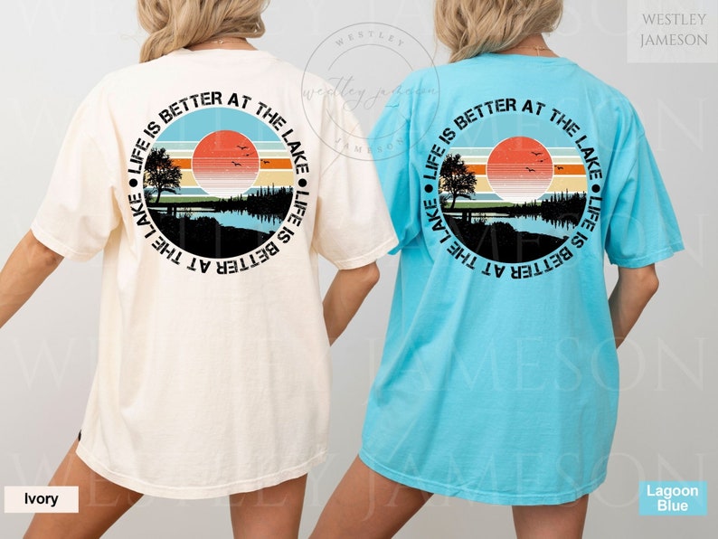 May include: Two t-shirts with a graphic design on the back. The design is a circle with a sunset, trees, and a lake. The text around the circle says "Life is better at the lake". The t-shirt on the left is ivory and the t-shirt on the right is light blue.