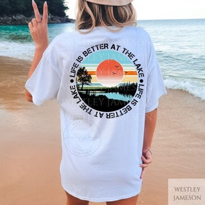 May include: White t-shirt with a circular graphic design featuring a sunset over a lake with a tree and the text "Life is better at the lake" in a black circle.