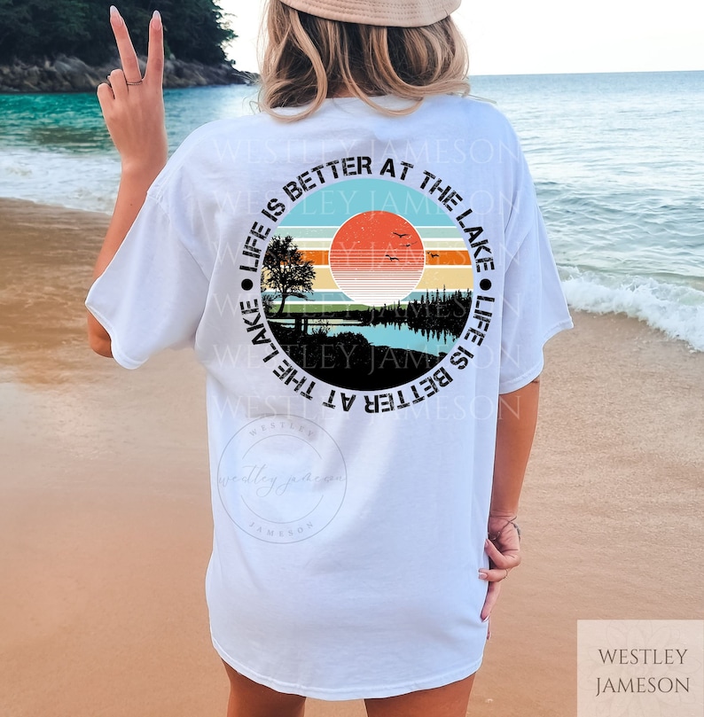 May include: White t-shirt with a circular graphic design featuring a sunset over a lake with a tree and the text "Life is better at the lake" in a black circle.