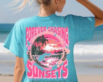 May include: A turquoise blue t-shirt with a pink and white graphic design of a sunset, palm trees, and waves. The text "FOREVER CHASING SUNSETS" is written in a pink and white font. The text "A GREAT SUNSET SURF AND PARADISE EVERY TIME LET'S GO TO THE BEACH" is written in a smaller font below the graphic.