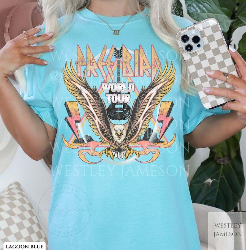 Pu&ograve; includere: Una t-shirt blu chiaro con una grafica di un'aquila con le ali spiegate, una chitarra e il testo "Free Bird World Tour" in stile vintage.