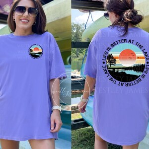 May include: A purple t-shirt with a graphic of a sunset over a lake and the text "Life is better at the lake" in a circle. The text "Westley Jameson" is printed on the back of the shirt.