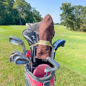 May include: A red and gray golf bag filled with golf clubs, including PING irons and various club head covers. A brown towel is secured with a gold and yellow band. The bag is on a green golf course.