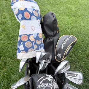 May include: A golf bag containing several metal golf clubs and protective headcovers. One headcover features a blue fabric with a repeating pattern of smiling cookies, milk cartons, and bananas. Other headcovers are black, including one labeled F-MAX.