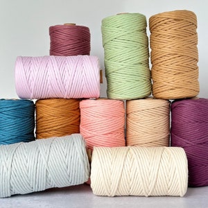 May include: A collection of spools of macrame cord in various colors, including teal, pink, and beige. The spools are arranged in a stacked display, showcasing the different shades and textures of the cord. The cords are tightly wound, ready for crafting projects.