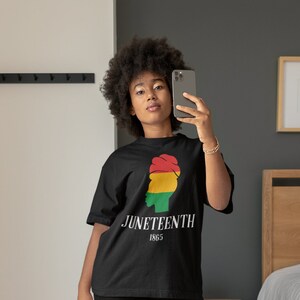 Op de afbeelding: Een persoon draagt een zwart T-shirt met een afbeelding van een vrouwenhoofd met een rood, geel en groen hoofddoek en de tekst "Juneteenth 1865". De persoon draagt een zwarte broek en witte sneakers.