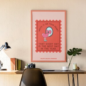 May include: A framed print with a pink background and a red stamp featuring a cartoon heart holding a mirror. The text on the stamp reads "Write it on your heart that every day is the best day in the year." The quote is attributed to Ralph Waldo Emerson.