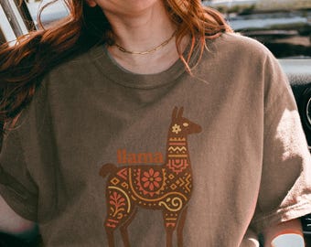 May include: Brown t-shirt featuring a decorative llama design with the text "Llama Queen" in orange. The llama has an intricate pattern in shades of brown and orange. The shirt is a neutral color.