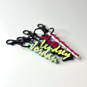 Puede incluir: Tres llaveros personalizados con cierres negros. Los llaveros presentan nombres en varios colores: "Jordan" en purpurina dorada, "Lindsey" en amarillo neón y un diseño rojo y blanco. Los llaveros están sobre un fondo blanco.