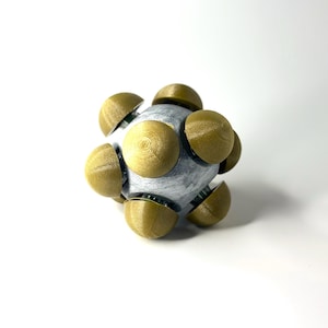 May include: A spherical object with a silver core and gold-colored, rounded segments arranged around its surface. The segments appear to be textured, and the object is set against a white background.