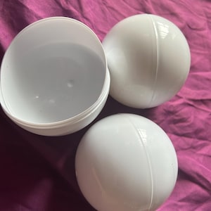 Puede incluir: Tres esferas de plástico blanco, una de ellas abierta para revelar su interior. Las esferas están sobre una superficie de tela morada. Las esferas son lisas y tienen una costura que recorre su circunferencia.