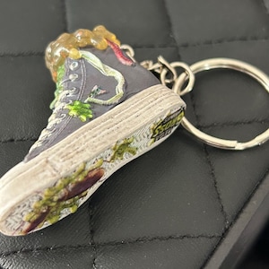 May include: A high-top sneaker keychain in dark gray with a distressed, textured sole. The shoe is adorned with green and brown embellishments, and a silver keyring is attached. The sole reads "SOSS LEGIT".