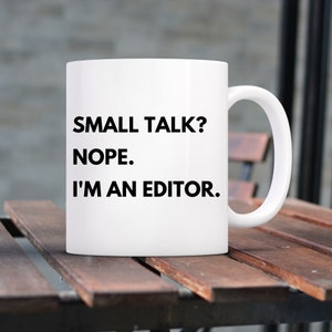 Peut inclure: Tasse en céramique blanche avec l'inscription noire : "SMALL TALK? NOPE. I'M AN EDITOR." Idéale pour un éditeur ou un écrivain. La tasse a une grande anse et est posée sur une surface en bois.