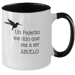 May include: White ceramic coffee mug with a black rim and handle. The mug has a black hummingbird silhouette and text that reads "Un Pajarito me dijo que vas a ser ABUELO".