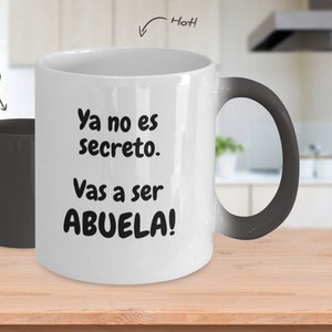 May include: A white ceramic mug with a black handle. The mug has a black heat-sensitive coating that reveals a message when hot: "Ya no es secreto. Vas a ser ABUELA!" The mug is on a wooden surface.