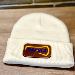 May include: White knitted beanie with a brown leather patch featuring the text "NAU" in blue and yellow lettering.