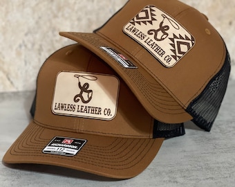Könnte beinhalten: Zwei braune Trucker-Caps mit schwarzen Netzrücken. Beide Caps haben einen braunen Leder-Patch mit dem Text "Lawless Leather Co." und einem Lasso-Design. Die obere Kappe hat ein geometrisches Muster auf dem Patch.