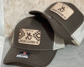 Könnte beinhalten: Zwei braune Trucker-Caps mit weißem Netzrücken. Beide Caps haben ein braunes Leder-Patch mit dem Text "Lawless Leather Co." und einem Lasso-Design. Das Patch auf der unteren Kappe hat ein indianisches Muster um das Lasso.