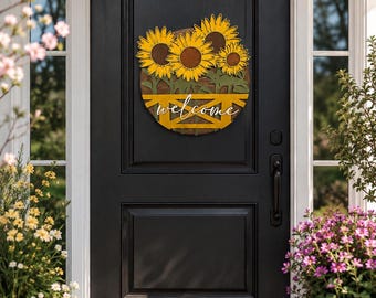 May include: A circular wooden door sign featuring three yellow sunflowers with dark brown centers above a yellow fence-style base. The word welcome is written in white cursive script across the fence. The sign is mounted on a black door.