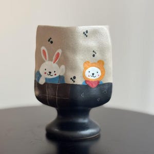 May include: A ceramic mug with a black and brown plaid design. The mug features a white rabbit and an orange bear wearing blue and red scarves. The mug has a black base.
