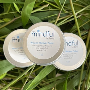 May include: Three round, silver-colored tins of "Wound Weaver Salve" are displayed on a bed of green leaves. Each tin has a white label with the text "the mindful herbalist" and product information. Each tin contains 1 oz of product.