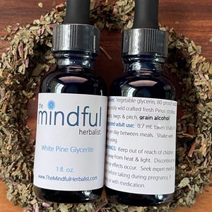 May include: Two 1-ounce bottles of White Pine Glycerite, a herbal remedy, labeled "The Mindful Herbalist". The bottles are dark blue with silver caps and droppers. The bottles are surrounded by dried herbs on a wooden surface.