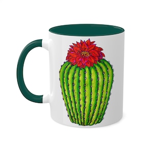 May include: A white ceramic mug with a green rim and handle. The mug features a hand-drawn illustration of a green cactus with a red flower on top.