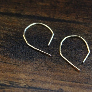 May include: A pair of delicate gold-colored hoop earrings. Each earring has a curved top and a straight, open bottom. The earrings are displayed on a dark brown wooden surface.