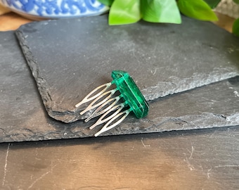 May include: A green rectangular hair comb with silver metal teeth. The comb is made of a translucent, emerald-colored material. The comb is resting on a dark gray, textured surface.
