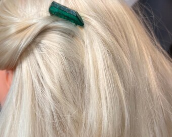 May include: A green crystal hair clip is inserted into blonde hair. The hair is styled in a bun. The crystal is a dark green color and has a geometric shape. The hair is long and straight.