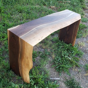 May include: A rustic wooden bench with a live edge top and a natural wood finish. The bench is made of walnut wood and has a unique, organic shape.