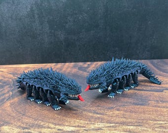 May include: Two black, spiky, insect-like figurines with red tongues. The figures have multiple legs and are posed on a wooden surface. The background is a dark, textured wall.