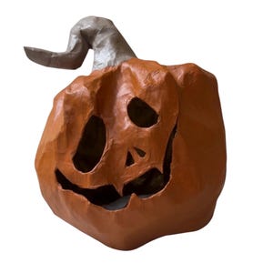 May include: A handcrafted orange pumpkin sculpture with a textured, paper-like finish. The pumpkin features a carved, asymmetrical face with hollowed-out eyes, a triangular nose, and a wide, jagged grin. A curved, light gray stem sits on top.