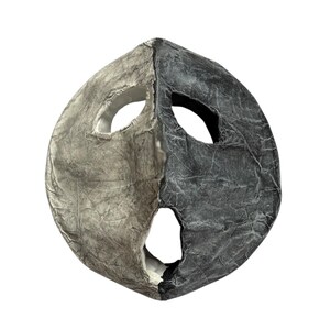 Peut inclure: Un masque fait à la main, divisé en deux parties, l'une gris clair et l'autre gris foncé. Il comporte des ouvertures pour les yeux et la bouche et a un aspect texturé, semblable à du papier. Le masque est de forme ronde.