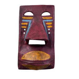 May include: A rectangular, maroon-colored mask with a carved face. The mask features two eye openings, a prominent nose, and a mouth opening. Yellow and blue painted accents adorn the face. The mask is likely for decorative purposes.