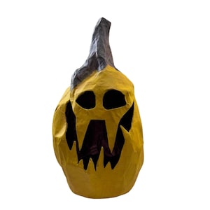 May include: A yellow papier-mâché pumpkin sculpture with a dark gray stem. The pumpkin features a carved, jagged face with two hollow oval eyes and a large, irregular mouth opening that reveals a dark interior, creating a textured, uneven surface.