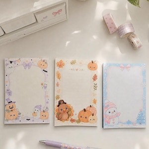 May include: Three rectangular memo pads featuring seasonal illustrations. The left pad shows ghosts and pumpkins, the center pad displays a turkey and autumn leaves, and the right pad depicts a snowman and winter motifs, all with pastel borders.