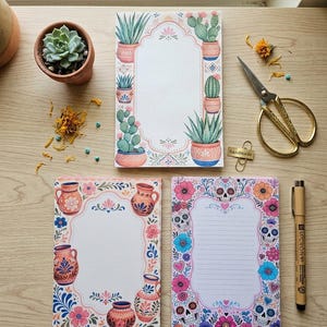 May include: Three rectangular stationery notepads featuring decorative borders. One notepad displays various potted cacti, another shows traditional clay pottery, and the third features sugar skulls and colorful flowers with lined paper.