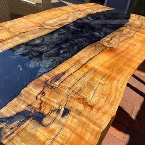 May include: A wooden table with a live edge and a black epoxy resin inlay. The wood has a natural grain pattern and is a light brown color. The epoxy resin is smooth and shiny.
