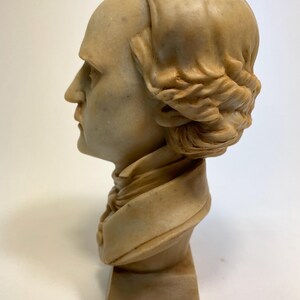 May include: A beige bust sculpture of a person with a braided hairstyle. The bust is on a beige pedestal.