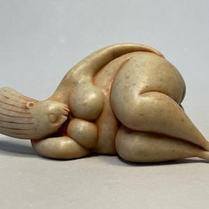 Puede incluir: Escultura beige de una figura reclinada. La figura está en posición fetal, con la cabeza apoyada en el brazo izquierdo. El cuerpo tiene formas redondeadas, con senos visibles y un abdomen prominente. La escultura tiene un acabado liso y mate.