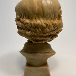 May include: A beige plaster bust of a person with curly hair. The bust is on a square base.