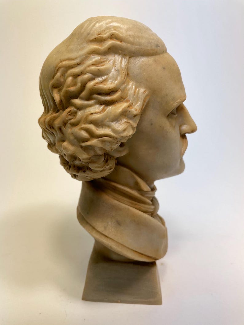May include: A beige plaster bust of a man with a sculpted hairstyle. The bust is on a square base.