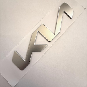 May include: A silver-coloured, stylised emblem with a geometric design, possibly a logo or badge. The emblem is on a clear adhesive backing, suggesting it's a sticker or decal. The design features sharp angles and a modern aesthetic.