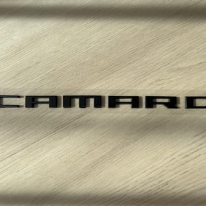 Peut inclure: Emblème CAMARO noir, accessoire automobile, présenté sur une surface en bois clair. Les lettres sont en gras et carrées, avec une finition mate. L'emblème est sur un support transparent.